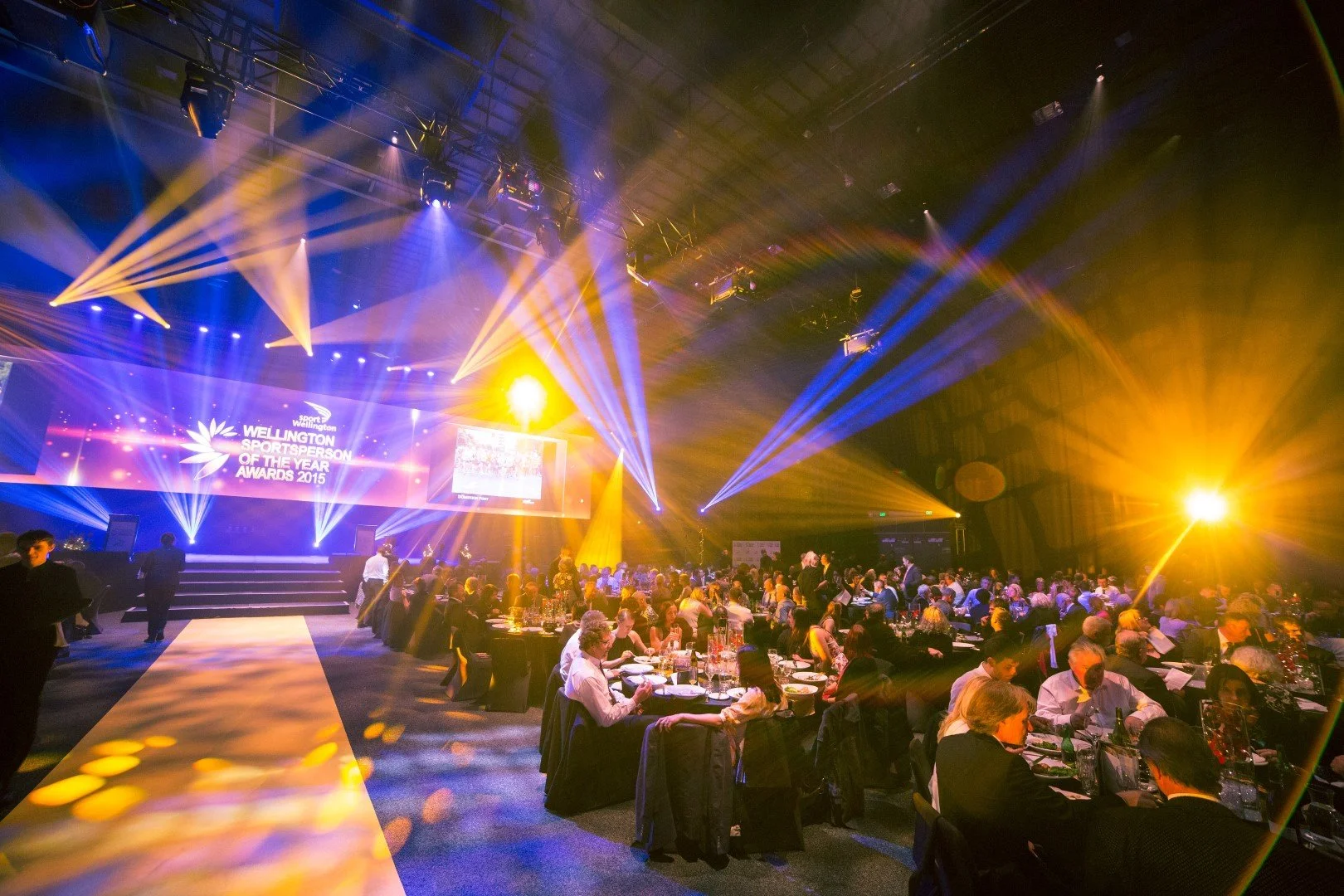 An awards ceremony with a stage and large screen displaying 'Wellington Sportsperson of the Year Awards 2015,' with bright yellow and blue stage lights illuminating the venue and guests seated at tables.