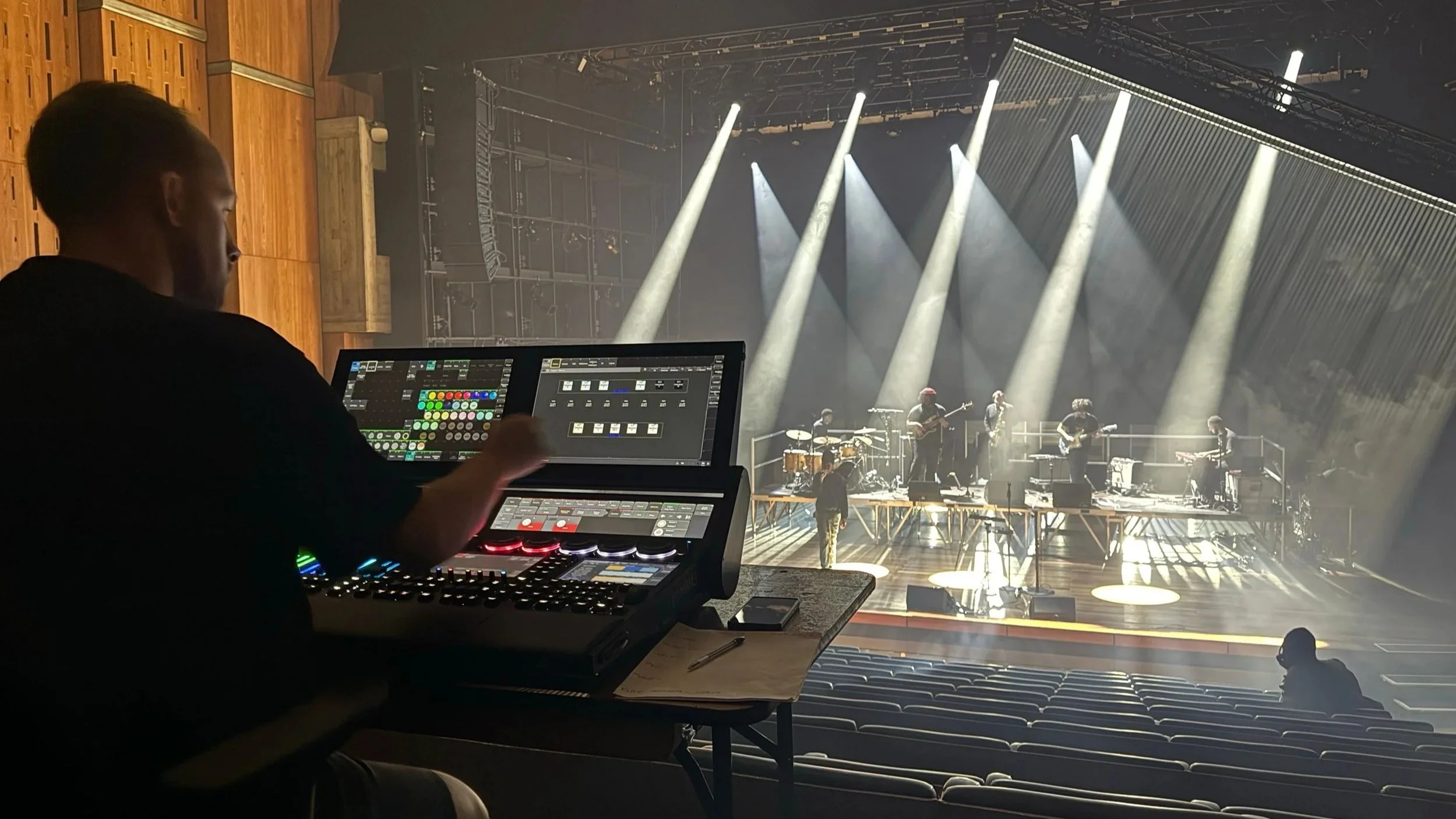 Lighting Designer working on a lighting console during a live band rehearsal on stage with musicians, bright stage lights, and empty audience seats.