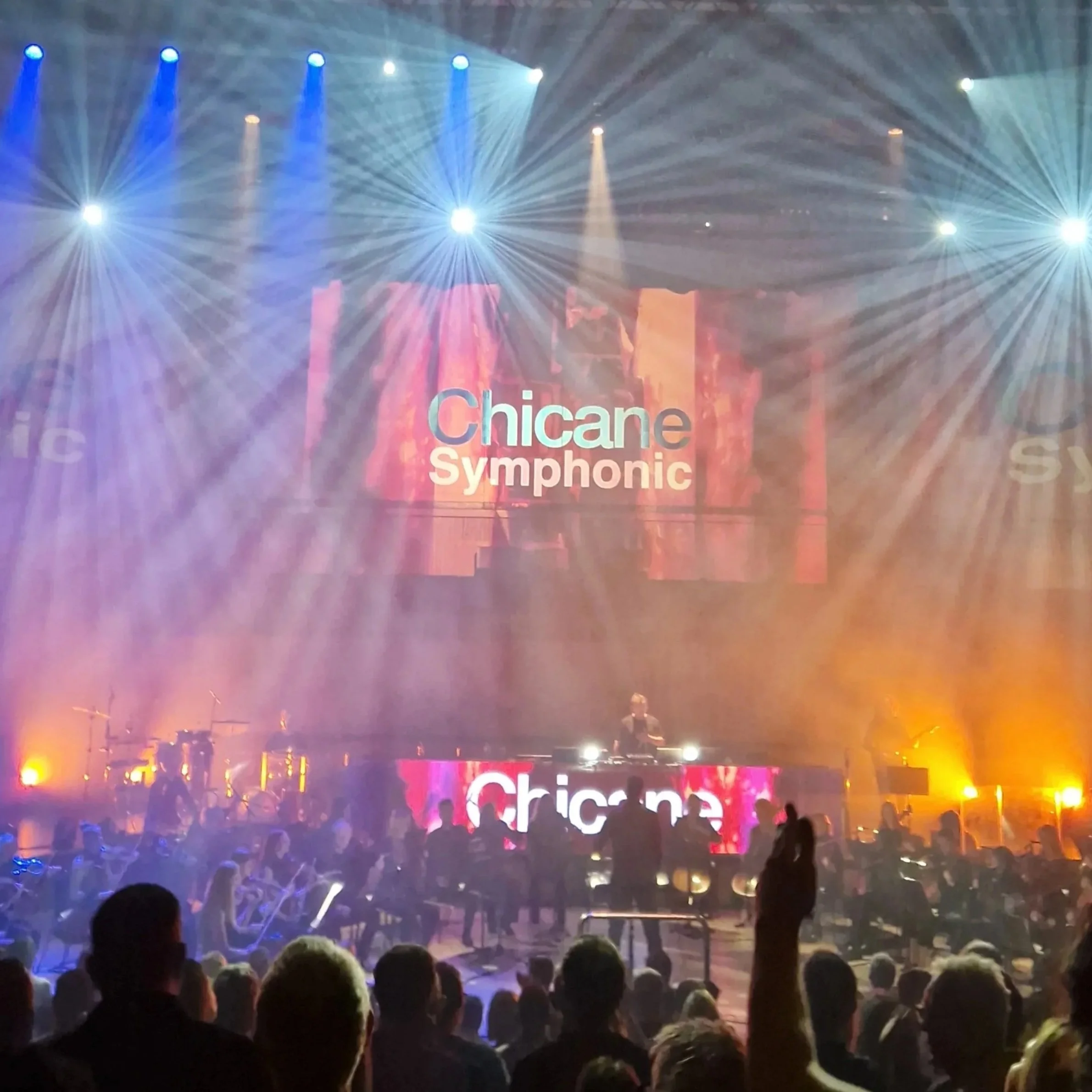 Concert scene with bright lights, a DJ at a console, and an audience, with the words "Chicane Symphonic" projected on a large screen.