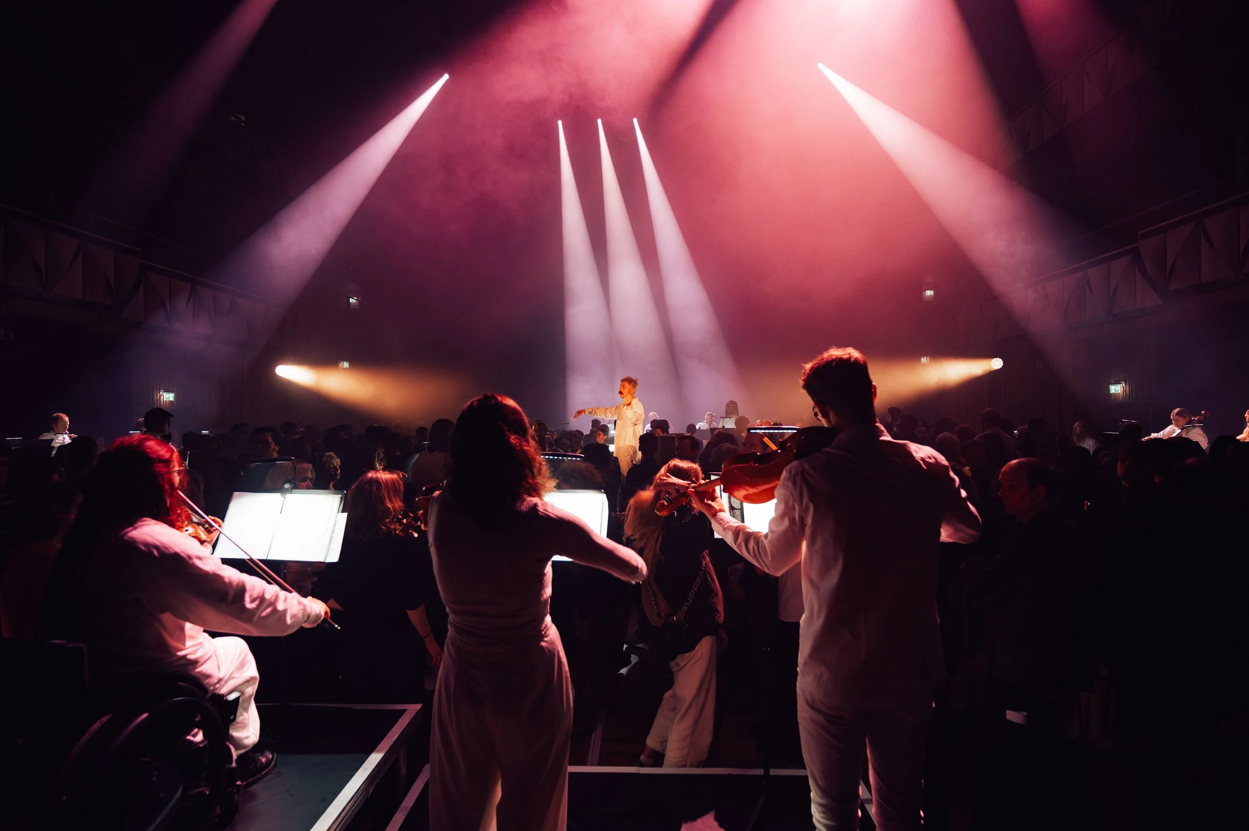 Concert with orchestra and conductor illuminated by pink and white stage lights in a dark theater.