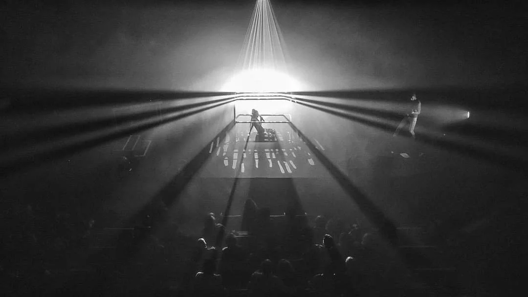 Black and white photo of a concert with intense lighting and shadows, with a performer on stage and a crowd watching.