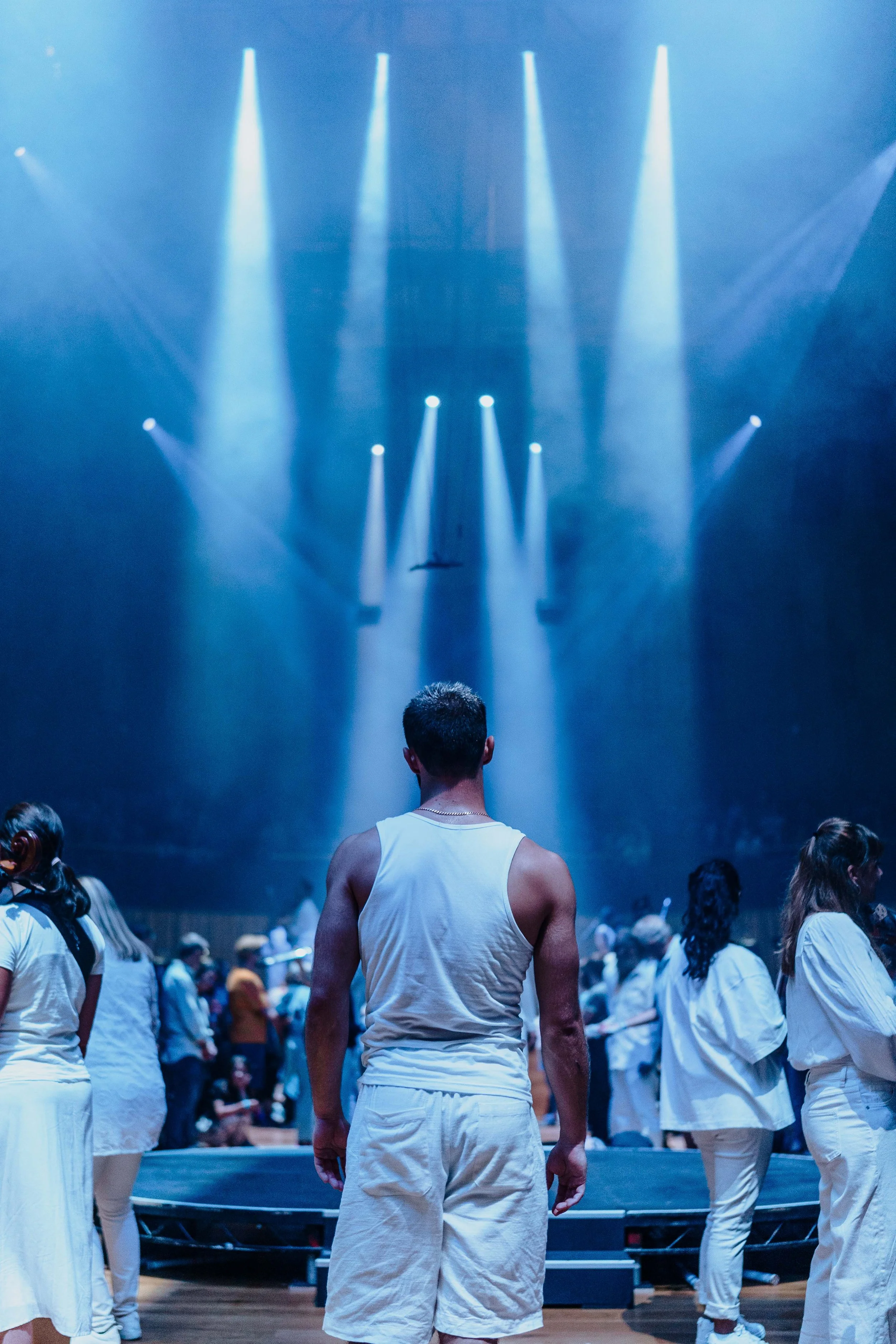 Performance with actors in white clothing on stage, dramatic lighting, and an audience in the background.