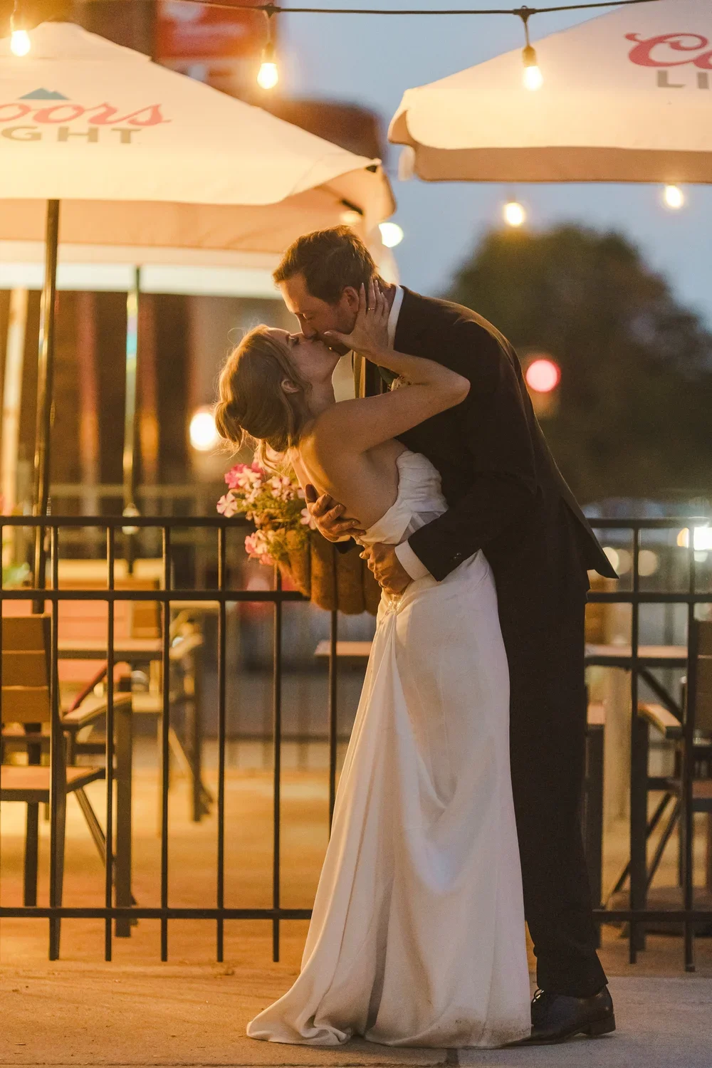 A couple sharing a romantic kiss at sunset or evening on an outdoor patio, with string lights overhead and umbrellas in the background, dressed in wedding attire.