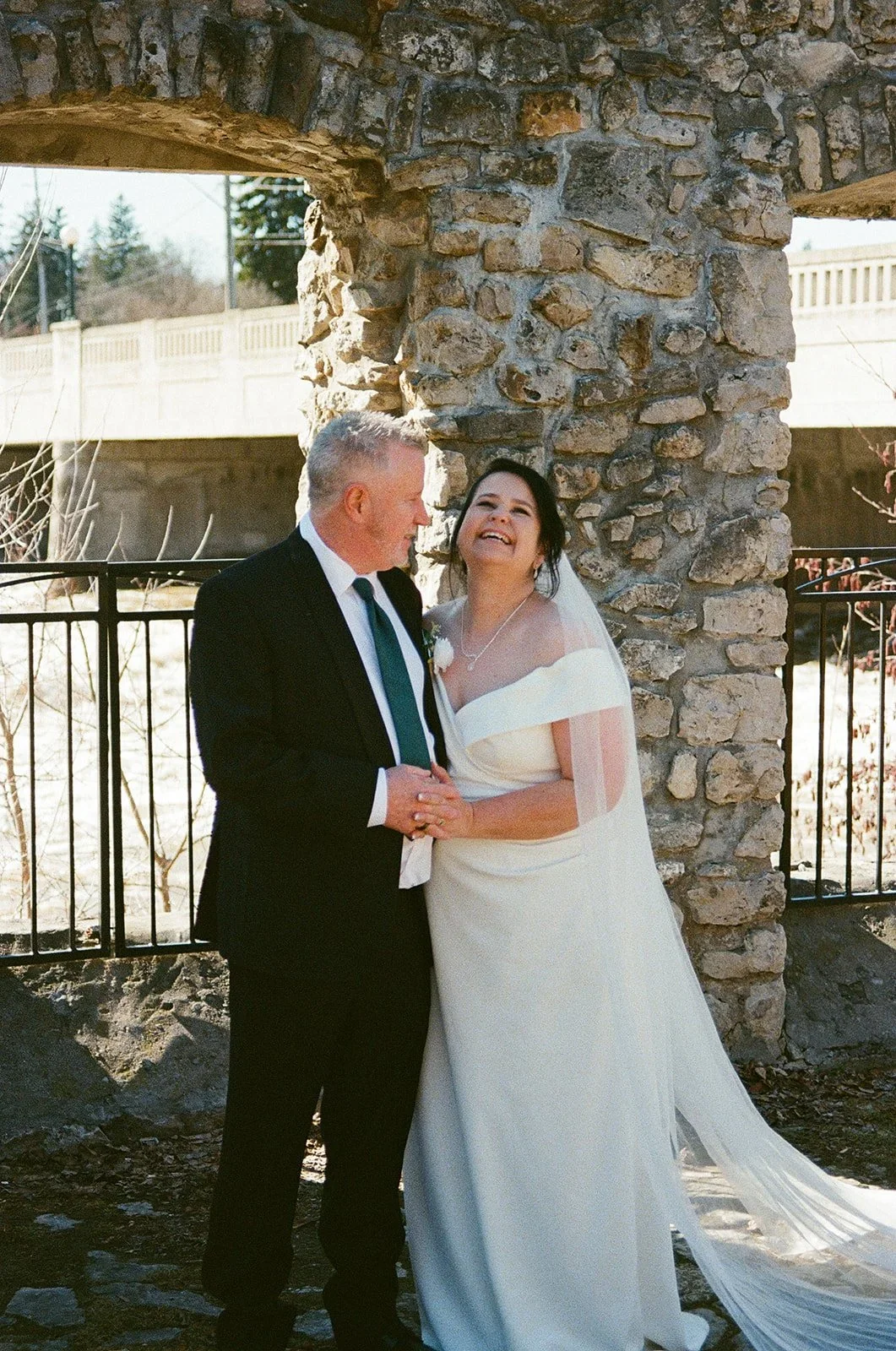 A bride and an older man, possibly her father, are happily holding hands and smiling at each other outdoors near a stone structure and a black metal fence. The bride is wearing a white wedding gown and veil. It is a sunny day.