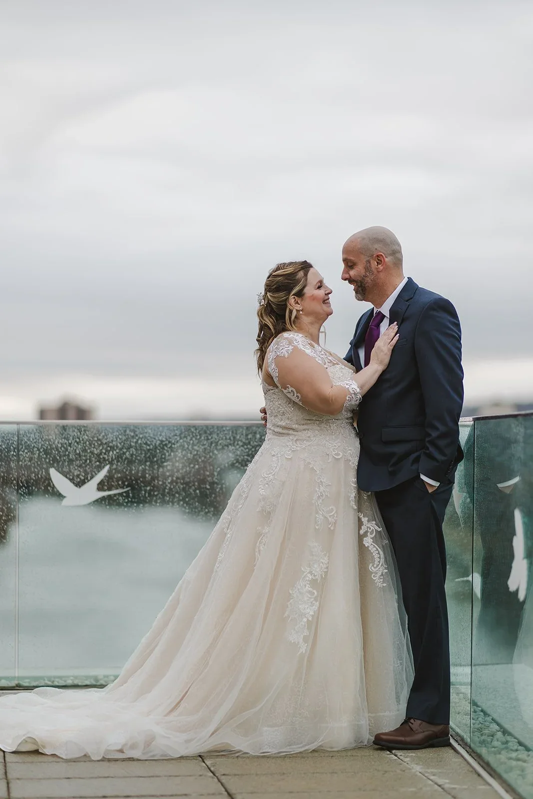 A bride and groom stand close together on a rooftop, smiling at each other against a cloudy sky with a glass railing behind them decorated with white bird silhouettes.