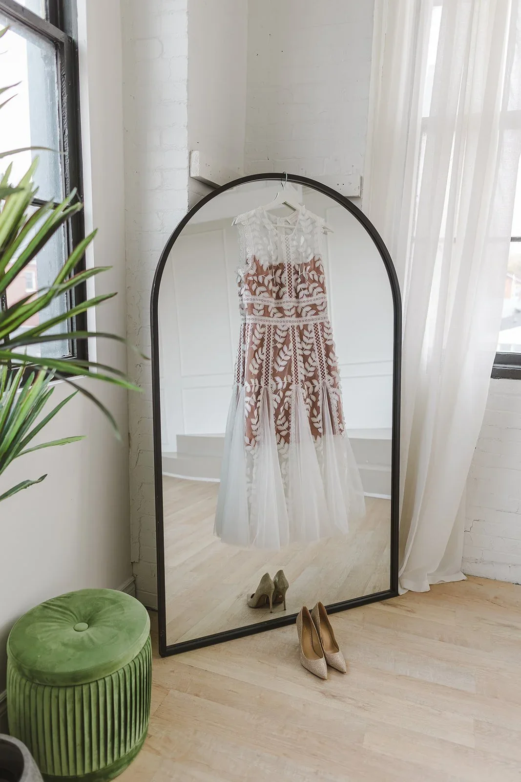 A full-length mirror reflecting a white and brown lace dress hanging on a wall, with a pair of beige high heels on the floor.