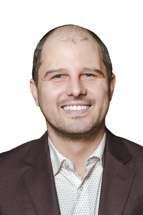 Headshot of a smiling man with short hair, wearing a dark blazer and white patterned shirt, against a white background.