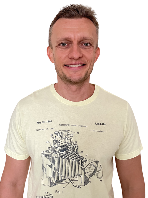A man with short brown hair and a slight beard, smiling, wearing a light-colored T-shirt with a vintage camera diagram printed on it.
