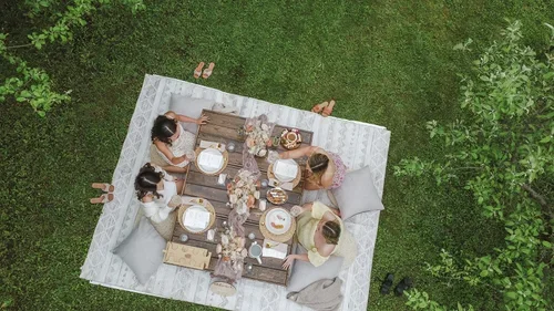 People having a meal outdoors on a large blanket in a grassy area with trees.