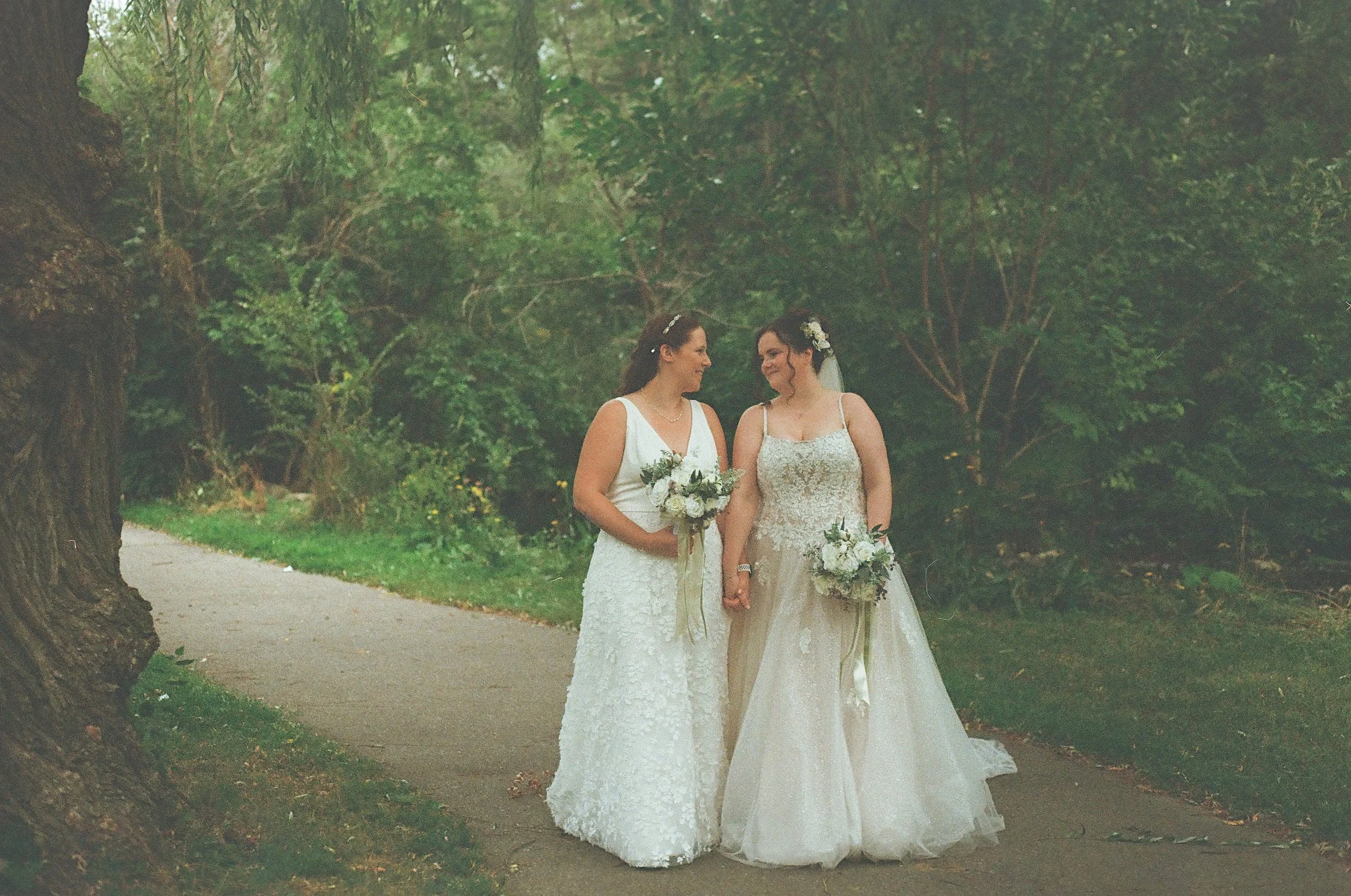 A couple getting married during an outdoor wedding ceremony at a wooden arch decorated with flowers, with an officiant in front of lush green trees and bushes.