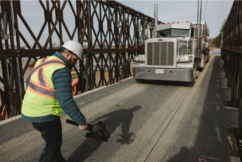 A construction worker in a reflective safety vest and white hard hat checks the surface of a steel bridge deck while a large semi-truck is parked on the bridge.