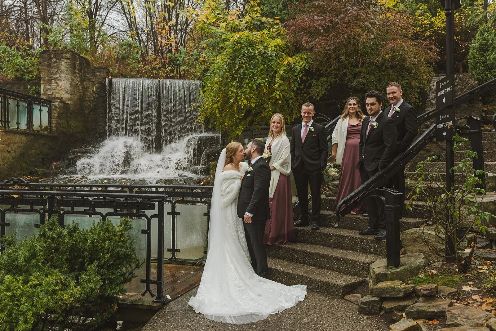 Bride and groom kiss by waterfall with wedding party on stairs  Ancaster,  ON  Ancaster Mill  Fedora Media.jpg