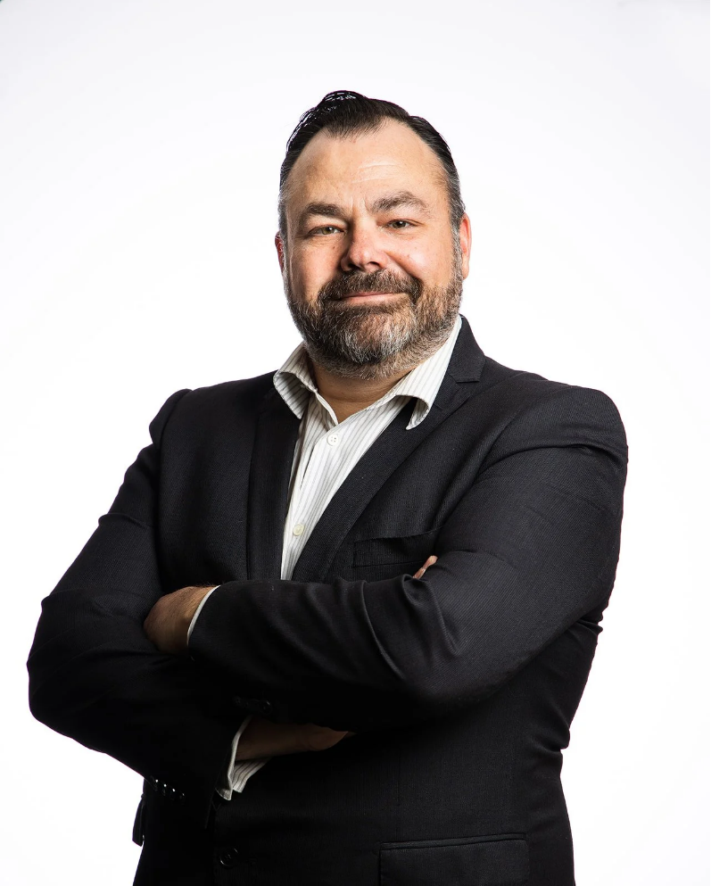 A man with dark hair and a beard wearing a dark suit jacket and white shirt, standing with arms crossed against a plain white background.
