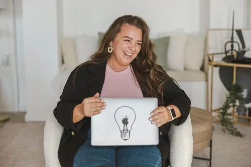 A woman sitting on a white chair, holding a laptop with a light bulb illustration, smiling and laughing in a modern living room.
