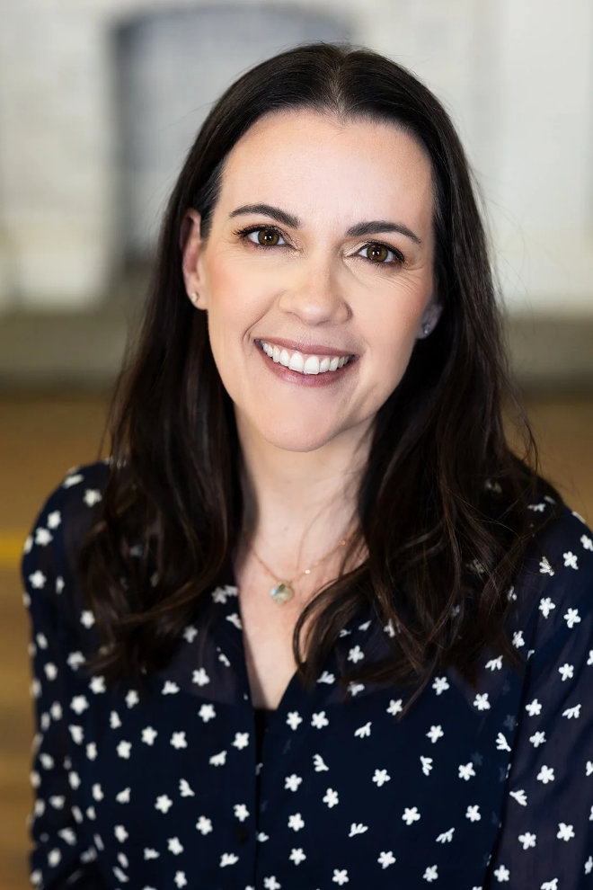 Headshot of a smiling woman with long dark hair, wearing a navy blouse with white floral pattern and a delicate necklace