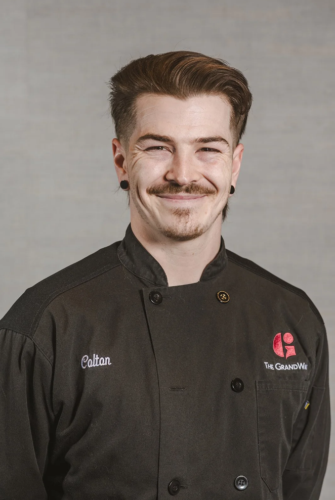 Portrait of a young man smiling, wearing a black chef's coat with embroidered name 'Calton' and a logo for The Grand W, against a neutral background.
