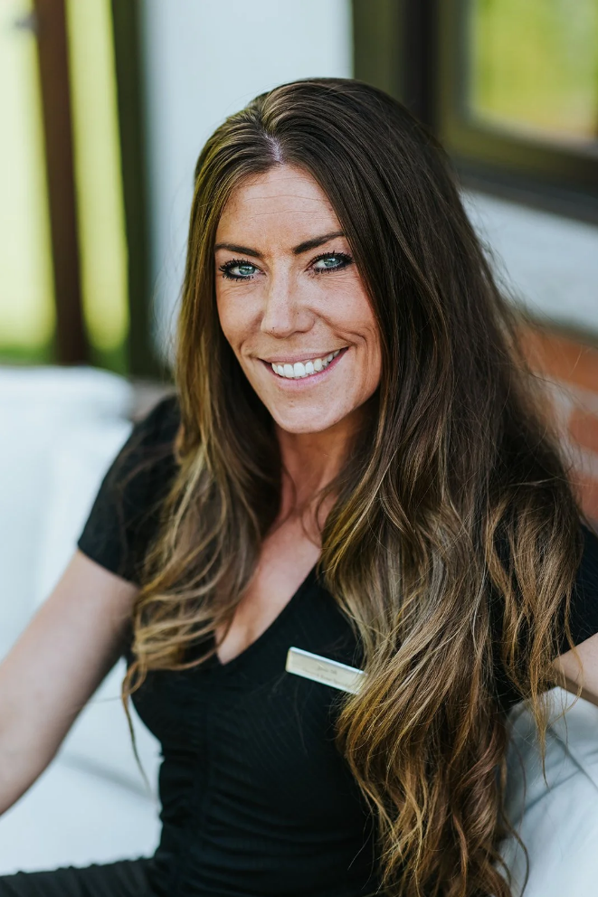 A woman with long, wavy brown hair and blue eyes, wearing a black top and a name tag, smiling while sitting indoors near a window with green scenery outside.