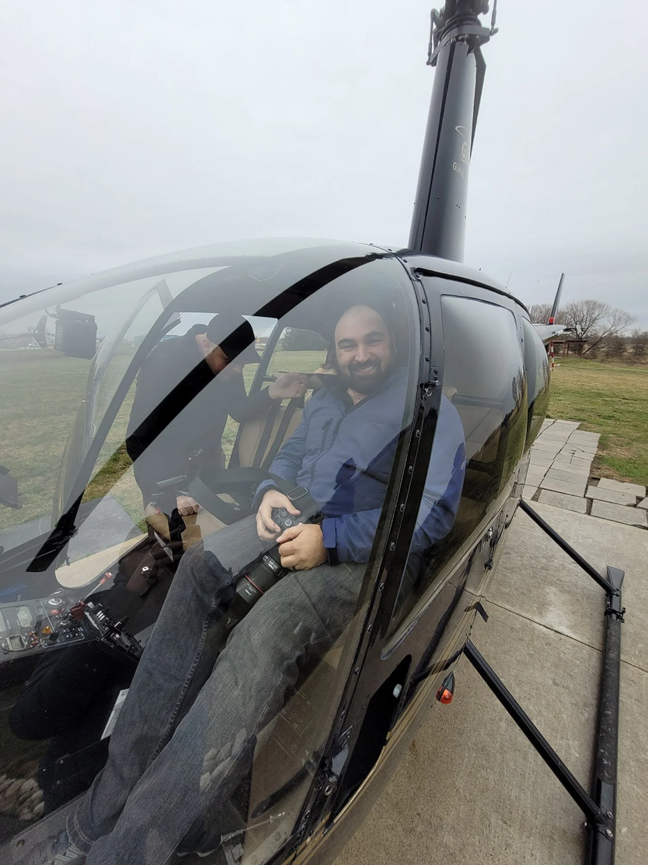 A man sitting inside a small helicopter with a clear canopy, smiling and holding a camera, on an outdoor tarmac with grassy fields and cloudy sky in the background.