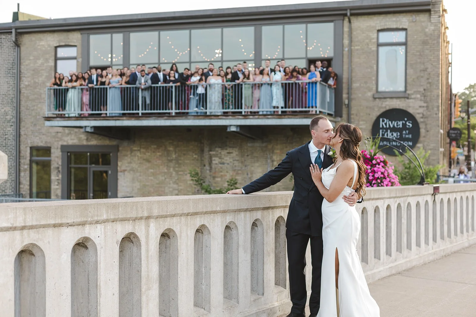 Bride and groom kissing on bridge with wedding guests behind on a balcony  River's Edge  Arlington Hotel  Paris, ON  Fedora Media.jpg