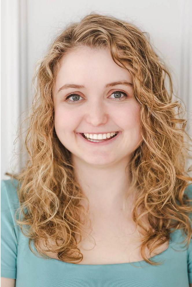 A young woman with curly red hair and blue eyes smiling at the camera, wearing a light blue top.
