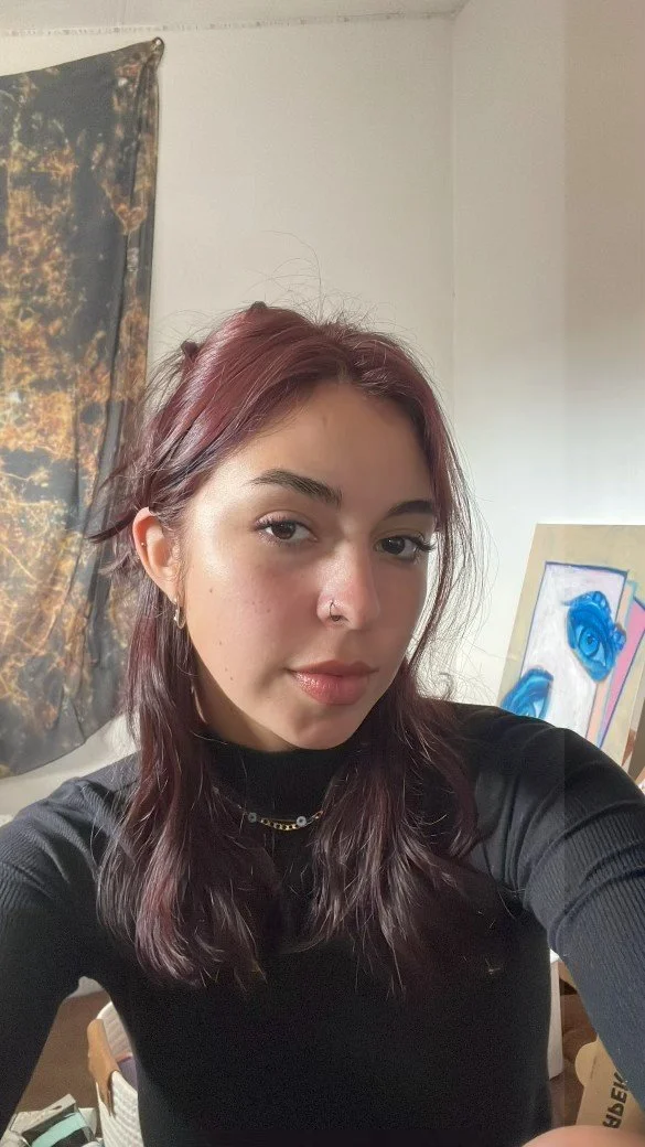 A young woman with shoulder-length dark red hair, wearing a black turtleneck and gold jewelry, taking a selfie in a room with abstract art and a dark patterned curtain in the background.