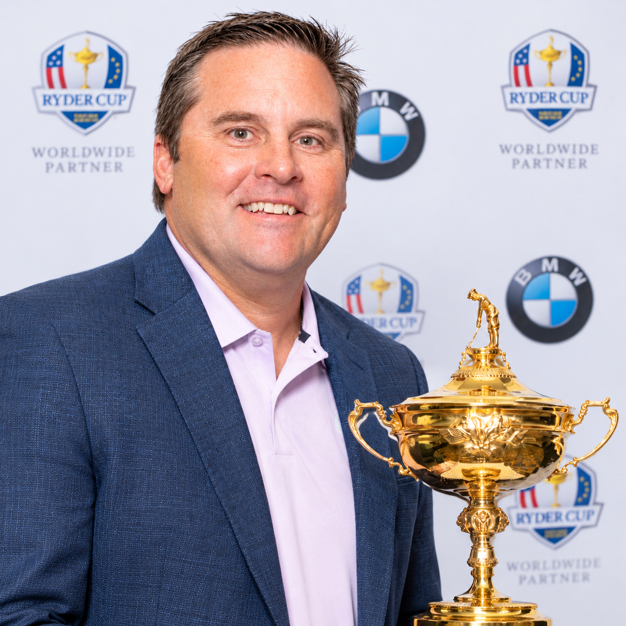 Smiling man in a blue blazer holding a gold golf trophy in front of Ryder Cup and BMW logos