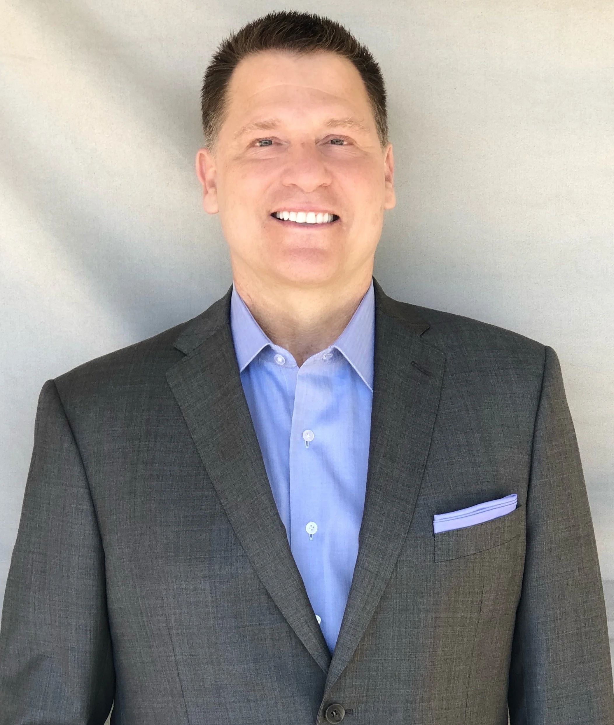 Close-up of a smiling man in a black suit jacket and light blue dress shirt, standing against a plain, light-colored wall.