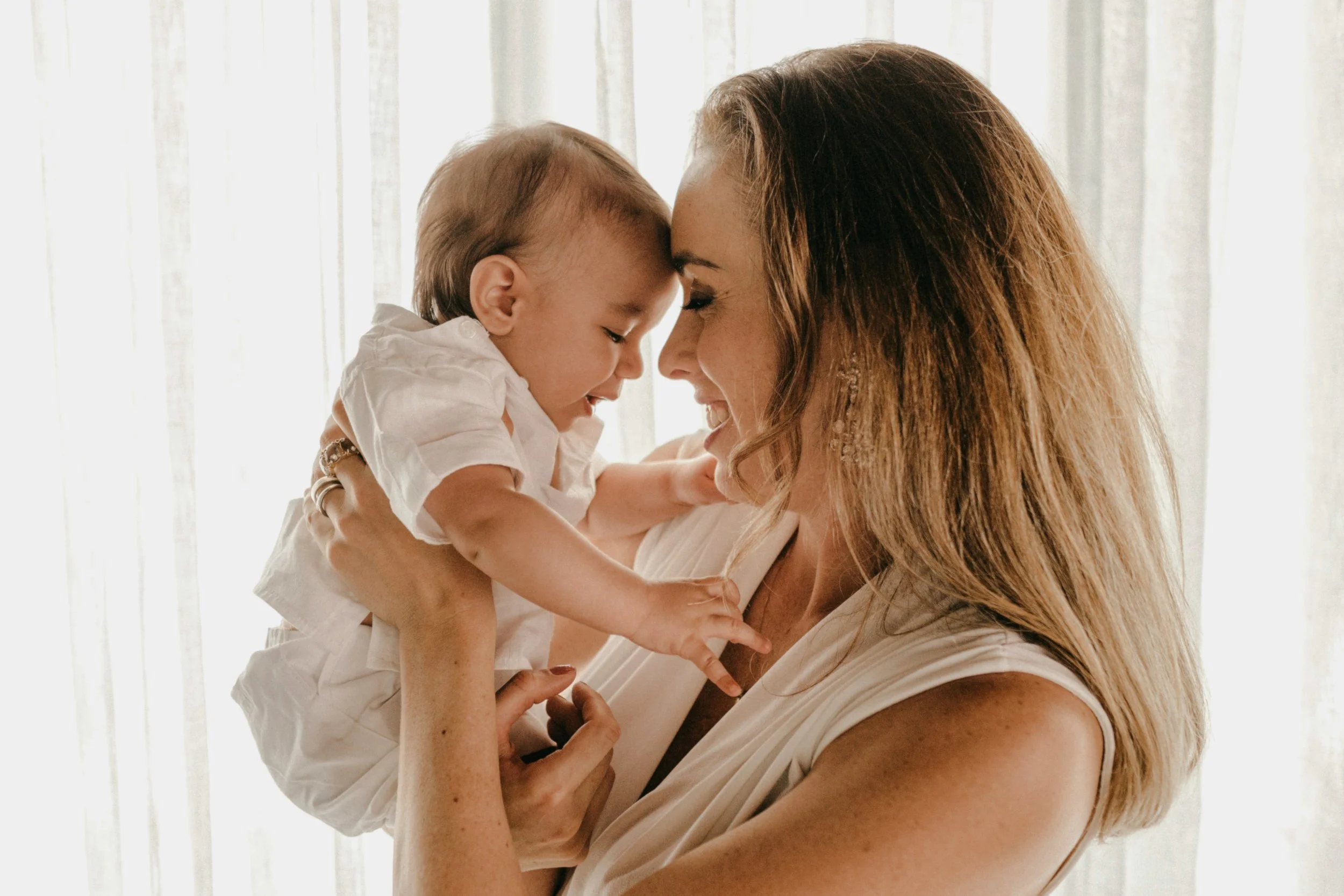 Une femme souriante tient un jeune enfant à bout de bras, leur tête se touchant, dans une pièce lumineuse avec des rideaux blancs en arrière-plan.
