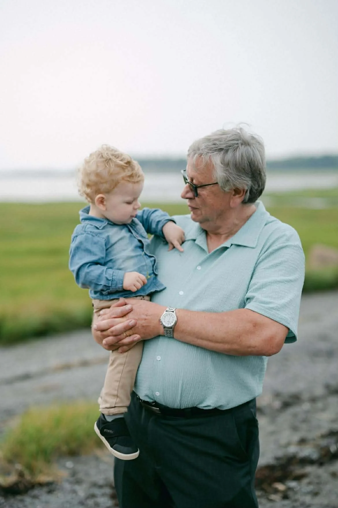 Un homme âgé tient un petit garçon dans ses bras, en plein air près d'un plan d'eau vert. Ils semblent échanger un regard ou une conversation, dans un environnement naturel.
