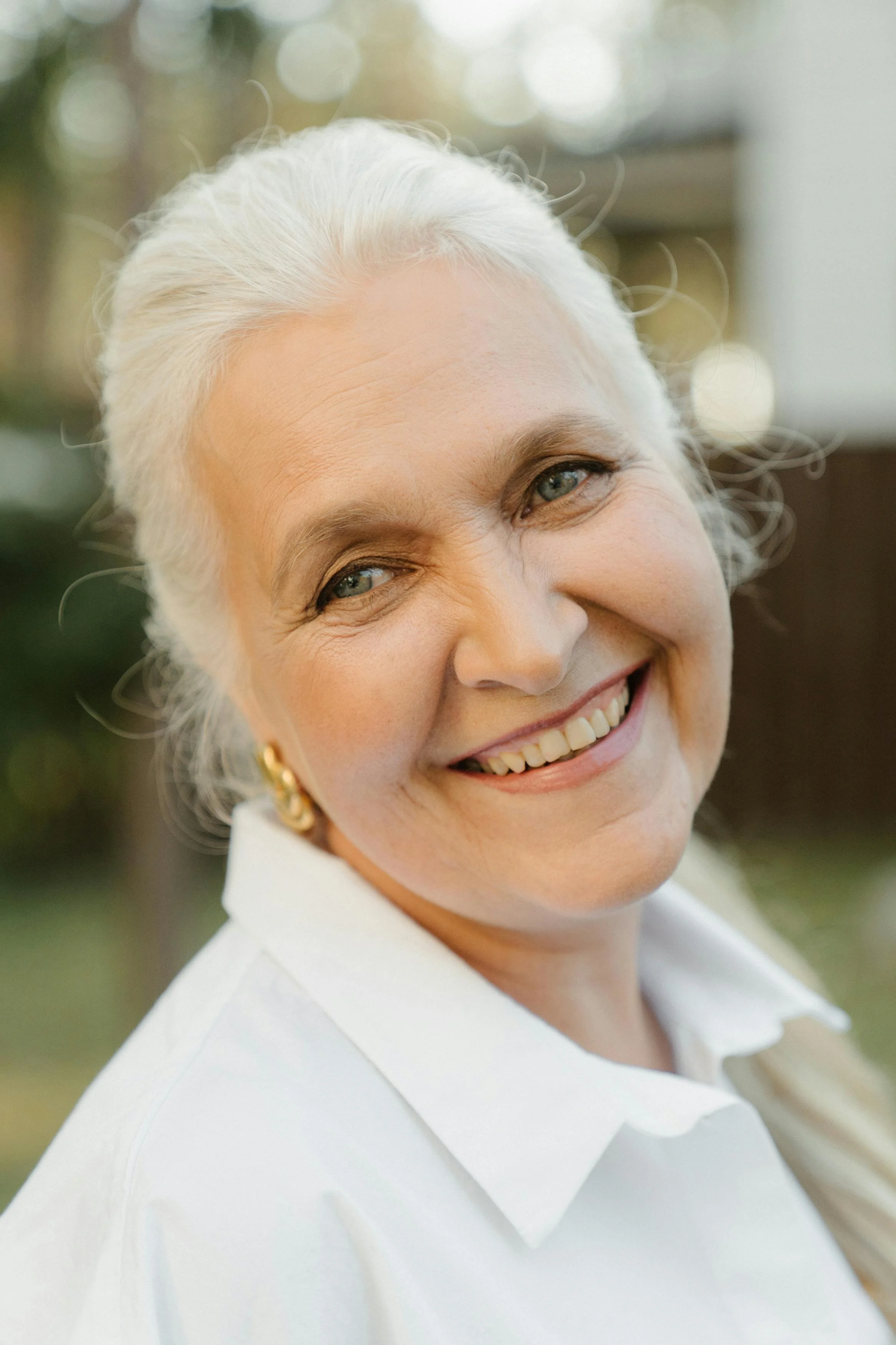 Une femme âgée souriante avec des cheveux gris bouclés, portant des boucles d'oreilles dorées et une chemise blanche, en extérieur avec un fond flou.