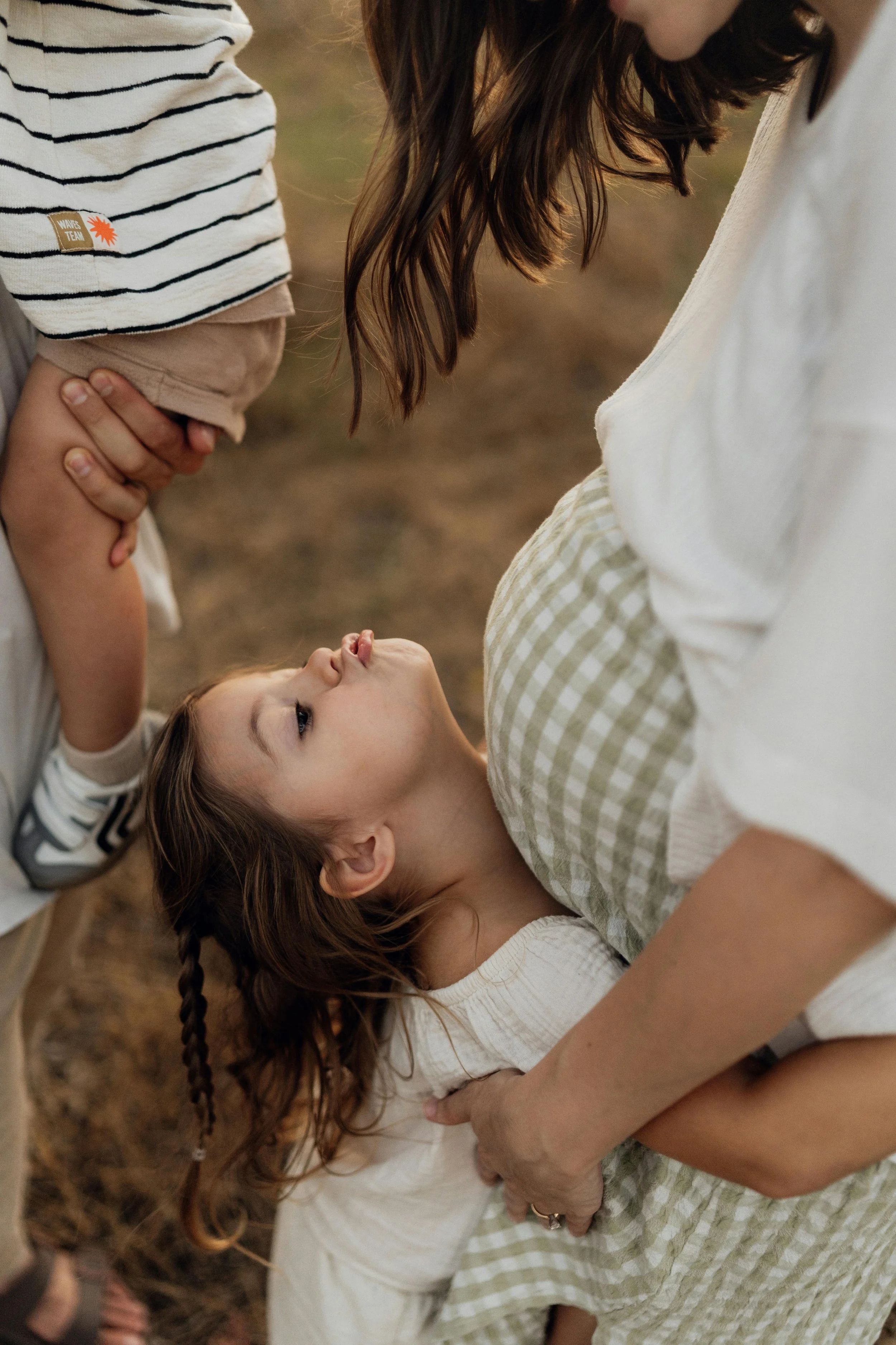 Une mère et ses deux enfants sur une plage, la mère regarde vers son enfant plus jeune qui se trouve dans ses bras, l'autre enfant est près d'eux, le tout dans une ambiance chaleureuse.