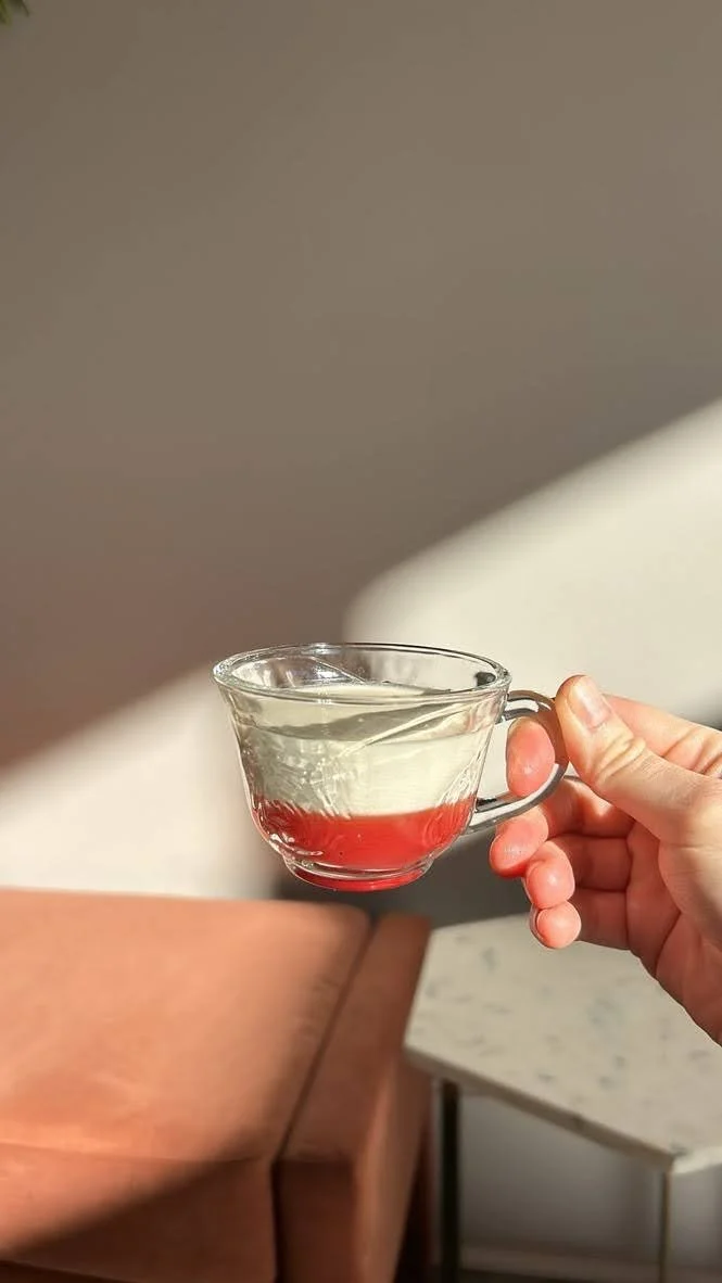 A person holding a clear glass teacup with a red liquid at the bottom, above a beige couch and a side table.