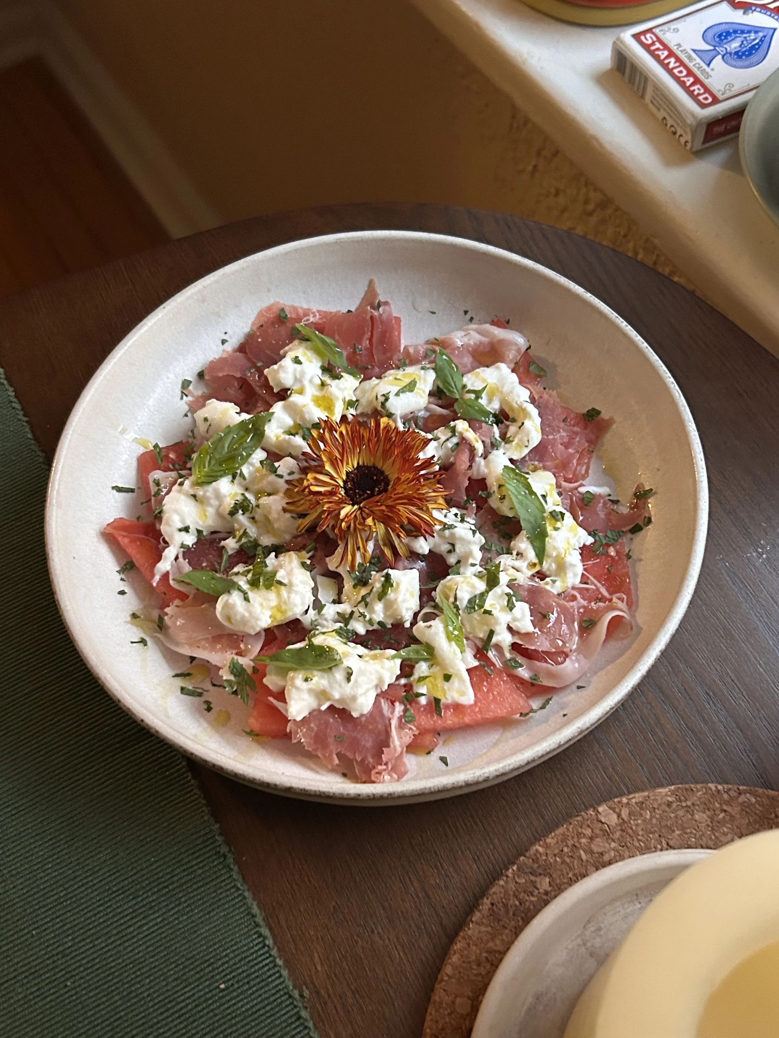 A plate of antipasto with sliced cured meats, crumbled cheese, fresh basil leaves, and a single edible flower on top.