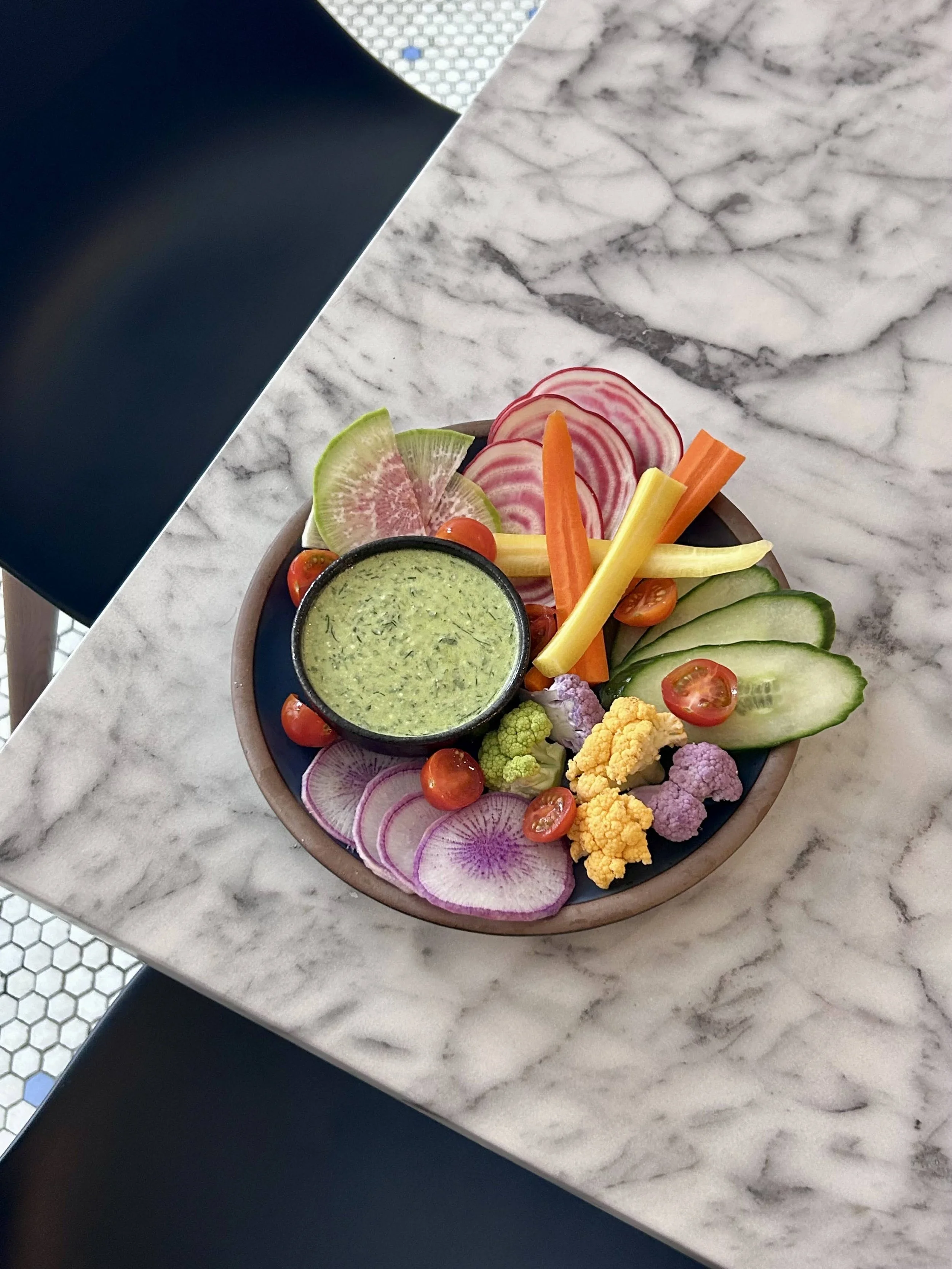 Colorful vegetable platter with slices of watermelon radish, cucumber, cherry tomatoes, cauliflower, carrots, and a small bowl of green dipping sauce, served on a dark plate on a white marble table.