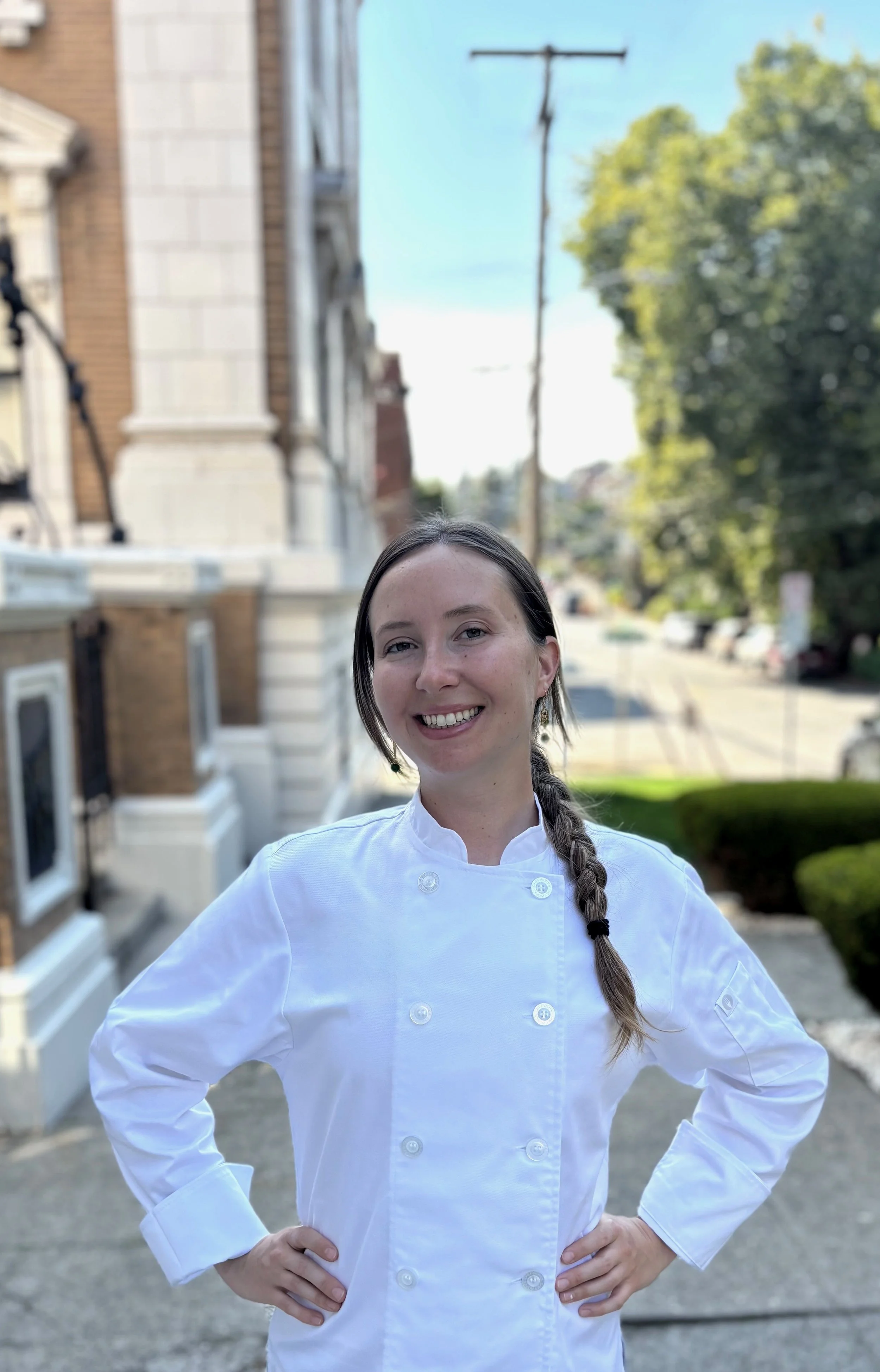 A woman wearing a white chef's coat standing outdoors with hands on hips, smiling, with trees, a street, parked cars, and a building in the background.