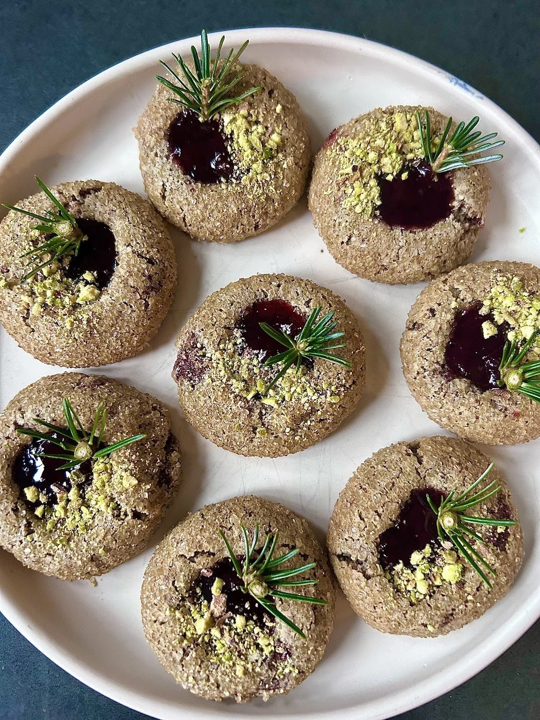 Plate of round cookies topped with cracked pistachios, dark jam filling, and sprigs of fresh rosemary.