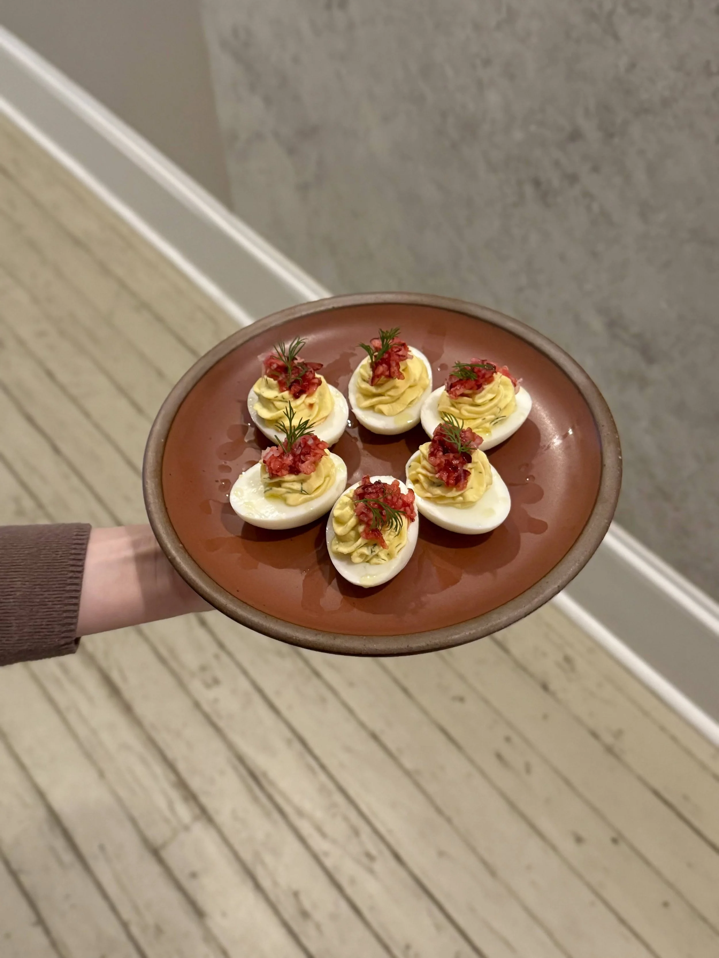 A person holding a brown plate with six deviled eggs garnished with herbs and red seasoning, set against a background of a wooden floor and a gray wall.