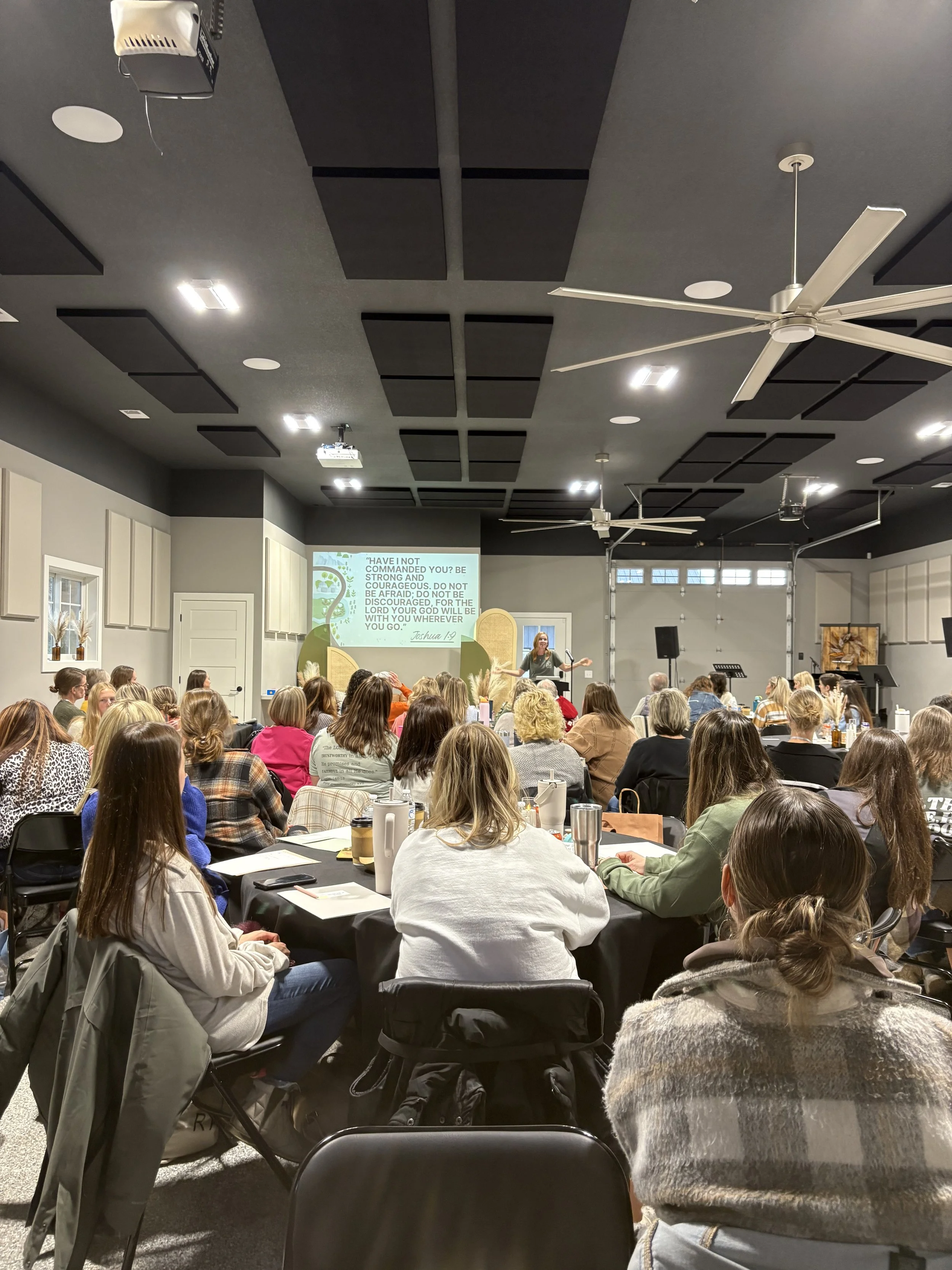 A woman standing at the front of a large room giving a presentation to an audience of mostly women sitting at tables. A slide with a Bible verse is projected on the wall behind her.