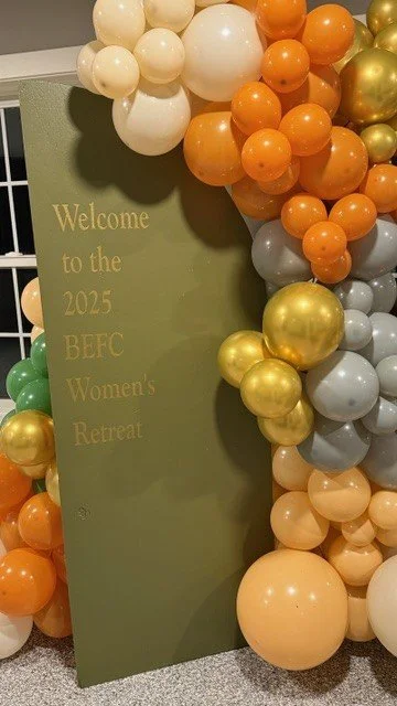 A green vertical sign that reads "Welcome to the 2025 BEFC Women's Retreat" surrounded by a colorful balloon arrangement in shades of white, orange, gold, green, and gray.
