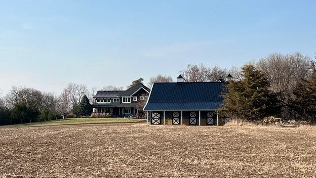 A farm scene with a large house in the background and a barn in the foreground, surrounded by leafless trees and open fields under a clear blue sky.