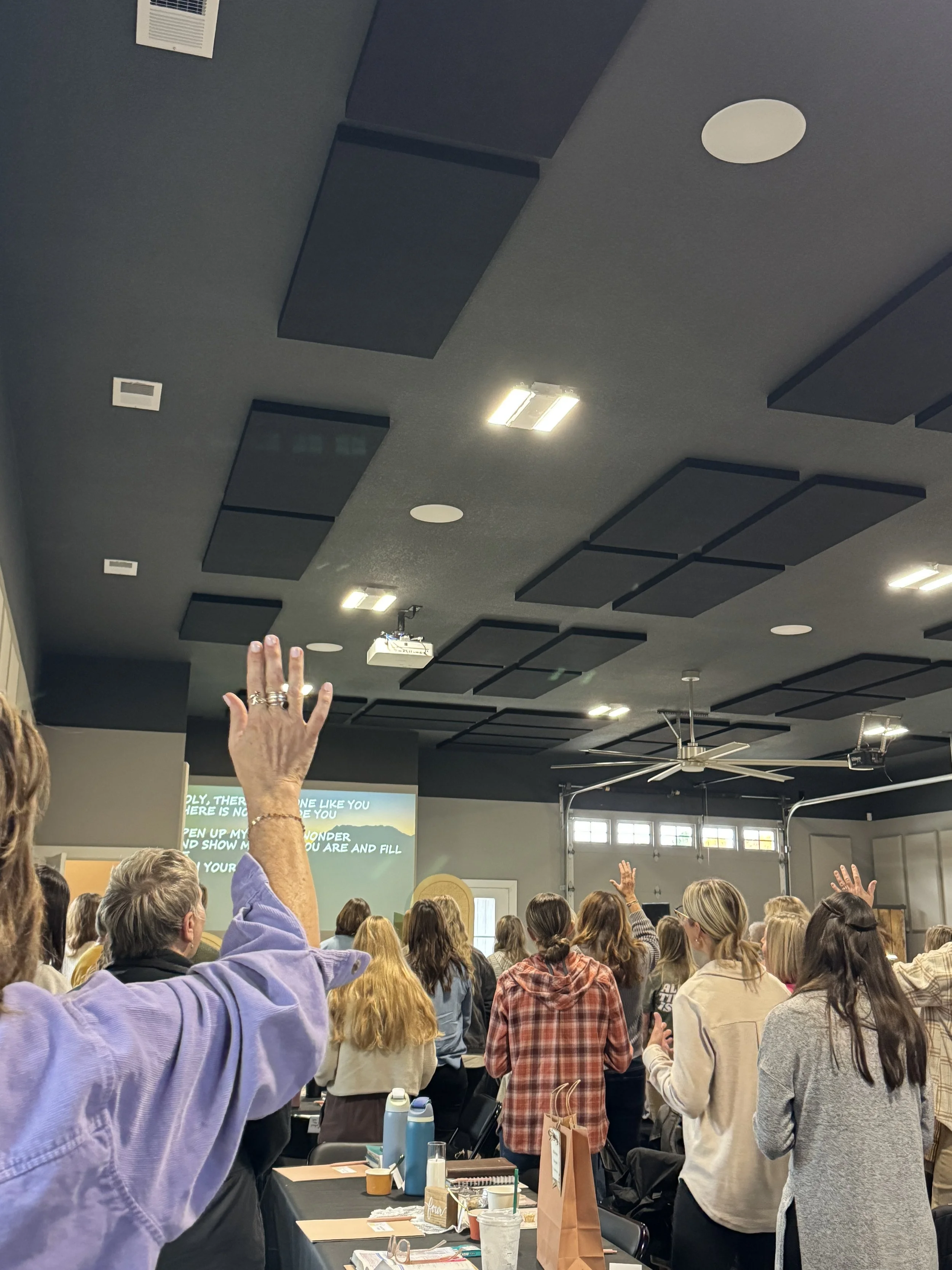 A group of people attending a conference. The room has a dark ceiling with acoustic panels, some ceiling lights, and a large fan. There is a projection screen showing text in the background.