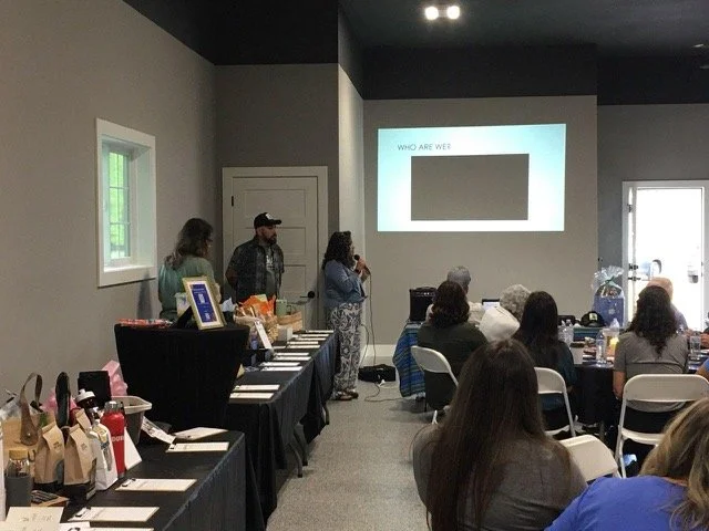 A woman speaking into a microphone at a conference with seated audience and a projected presentation on the wall.