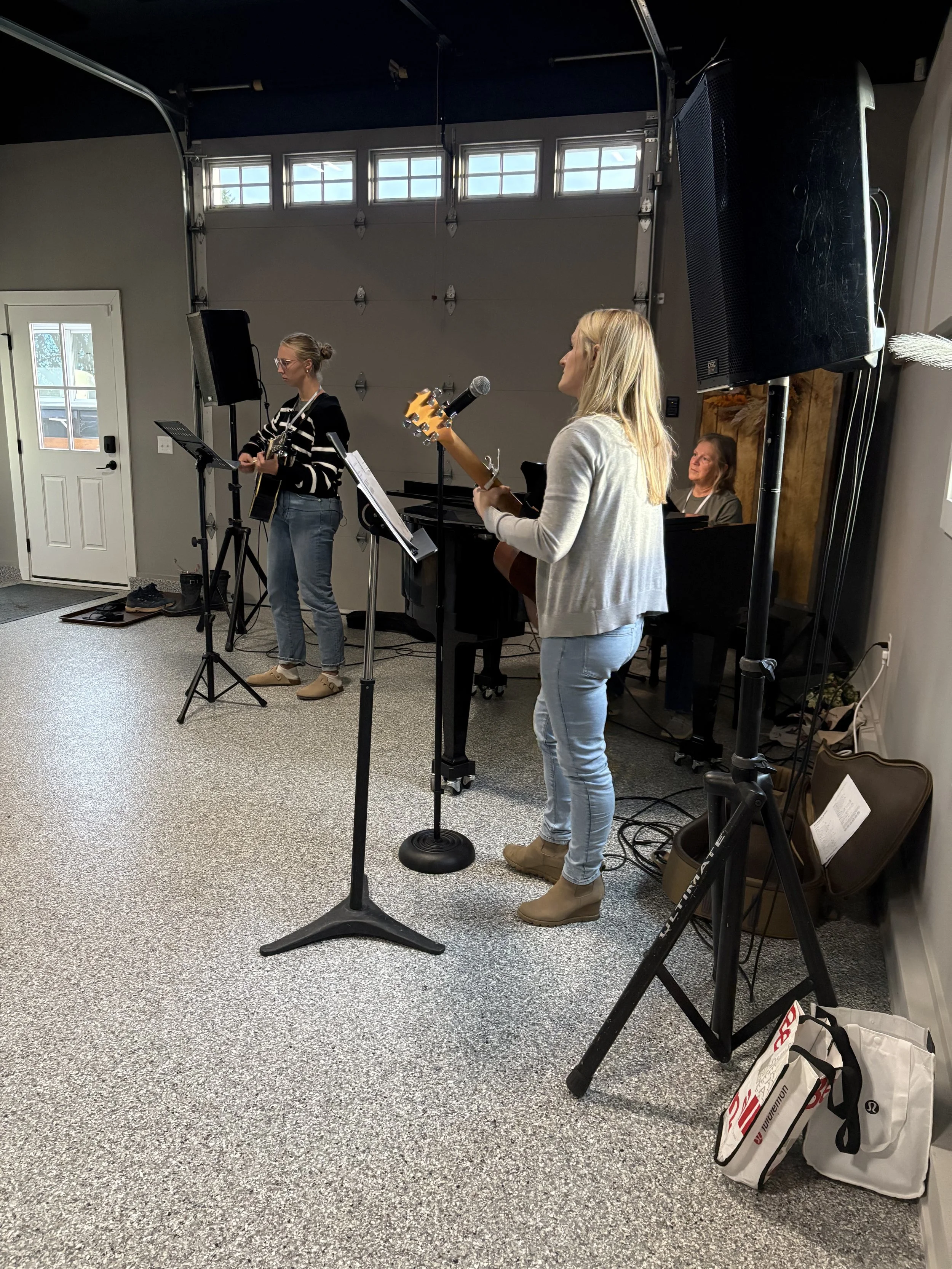 Two women playing guitars and singing into microphones in a room with a grey garage door and a piano in the background.