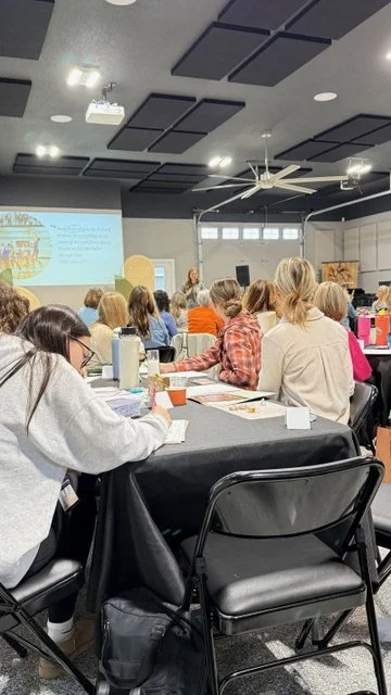 People attending a workshop with a presenter at the front, a projected presentation on the wall, and notebooks on the tables.