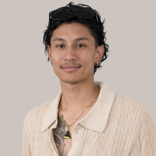 Portrait of a young man with dark, wavy hair, wearing a beige sweater, black sunglasses on his head, a necklace, and earrings, smiling at the camera against a plain background.