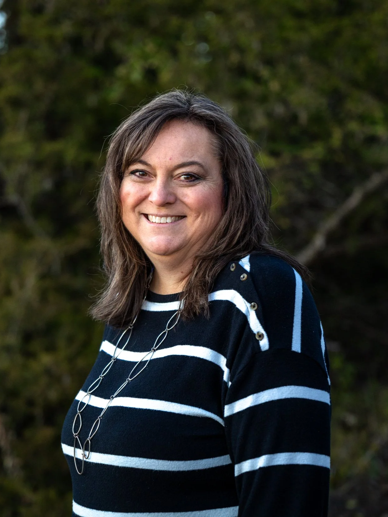 A woman with shoulder-length brown hair smiling outdoors, wearing a black and white striped sweater and a long silver necklace, with trees in the background.