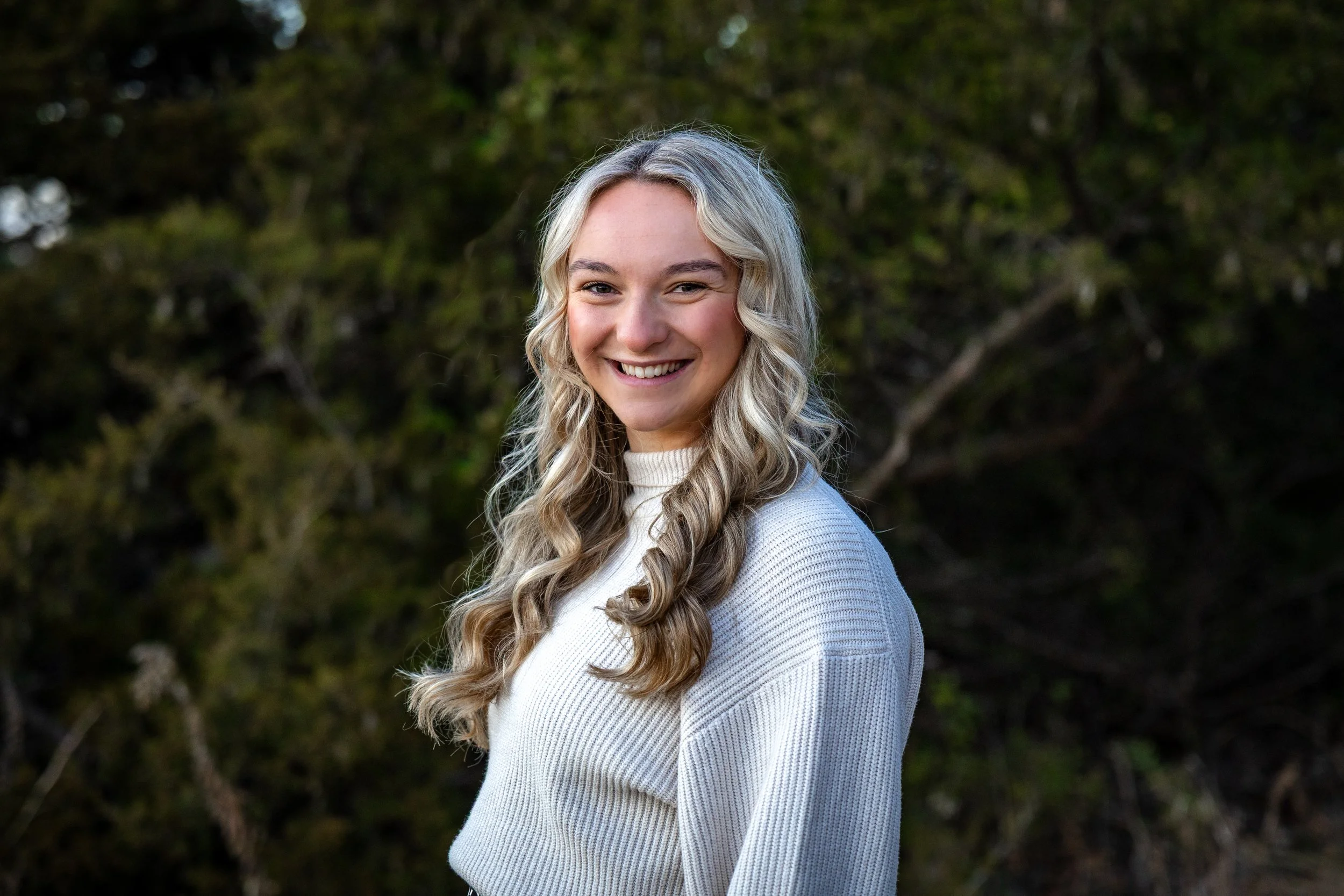 A smiling woman with long, wavy blonde hair standing outdoors against a background of green foliage.