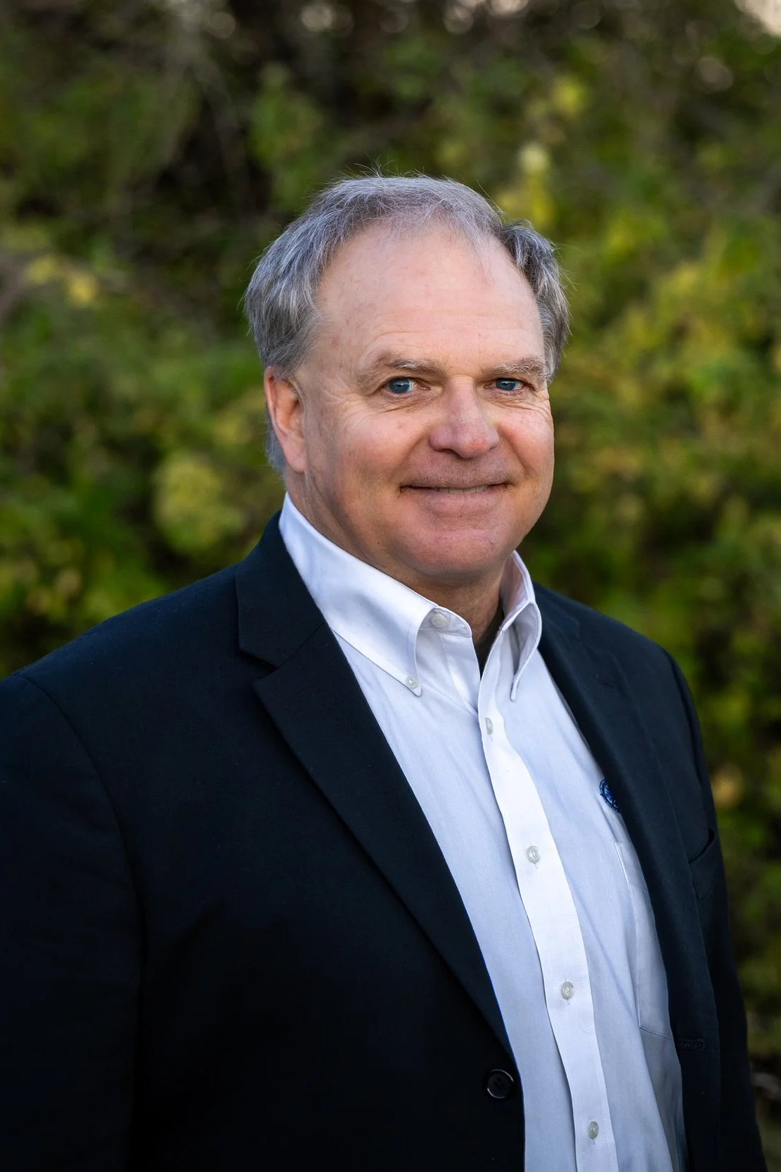 A middle-aged man with gray hair and blue eyes is wearing a white button-up shirt and a black blazer. He is standing outdoors with blurred green foliage in the background, smiling slightly at the camera.