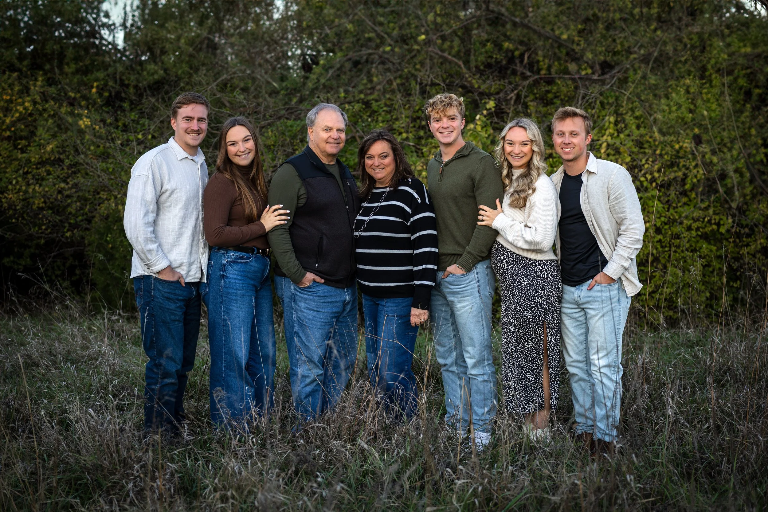 A family of seven standing outdoors in a grassy field with trees in the background