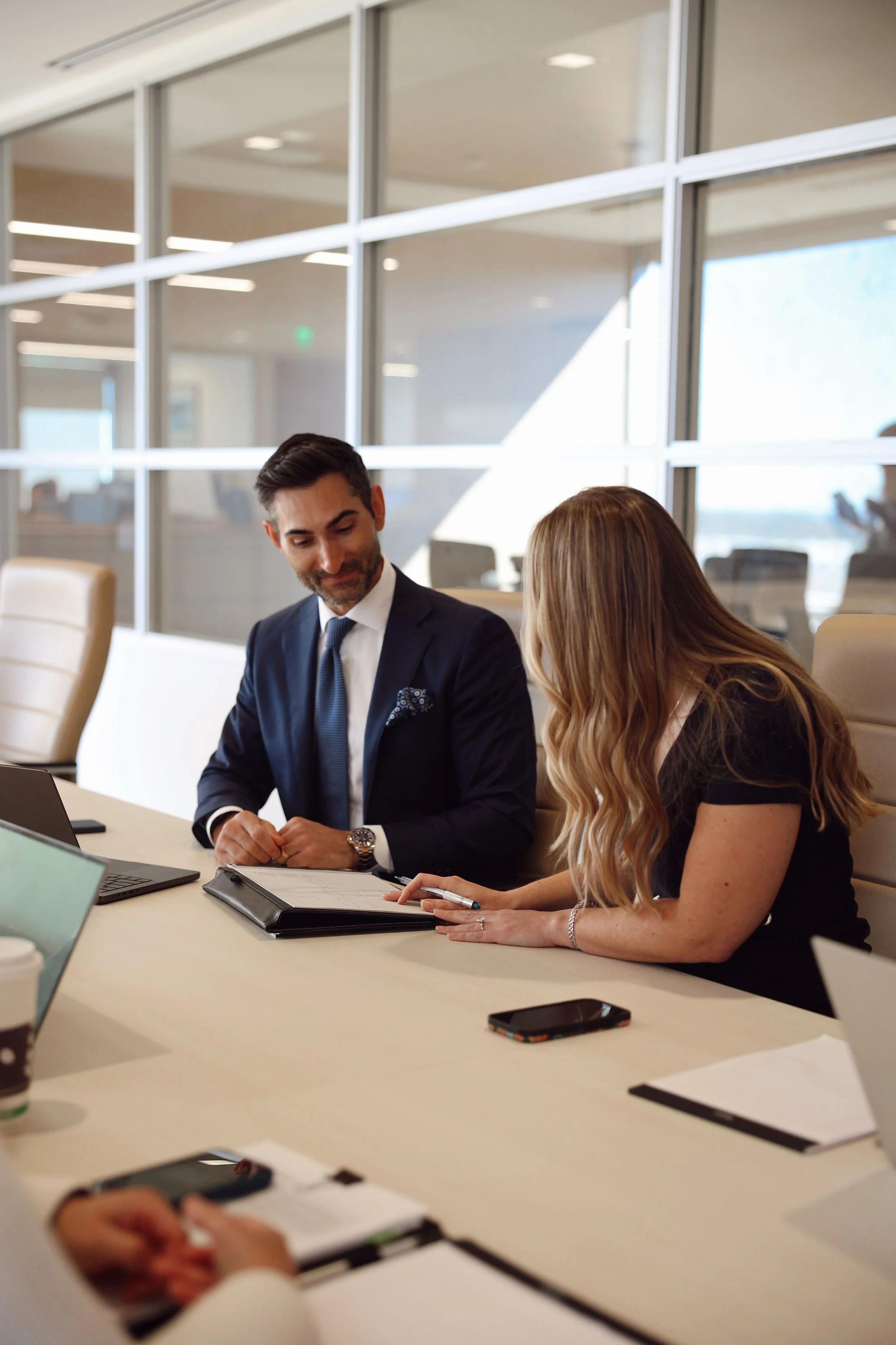 Two professionals, a man in a suit and a woman with long blond hair, are engaged in a discussion at a conference table in a modern office with glass walls, plants, and natural lighting.